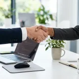 清潔な白いデスクで交わされる力強い握手。背景にはノートパソコンが置かれ、責任と約束（コミット）を表現している。明るい自然光のプロフェッショナルな写真。
