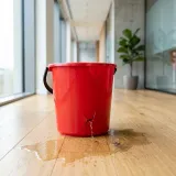 スタイリッシュなオフィスの床に置かれた、底が割れて大きな穴が空いている真っ赤なバケツ。そこから水がポタポタと床に漏れ出ている様子で、顧客がどんどん解約して流出していく「チャーン（解約）」の恐ろしさを象徴するイメージ
