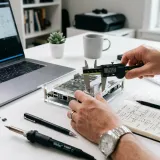 清潔な白いデスクに置かれたノートパソコン。その横では、人が専門的な道具を使いこなし、図面が描かれたノートに何かを書き込んでおり、長年の経験（ノウハウ）を表現している。