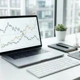 清潔な白いデスクに置かれたノートパソコン。その横には、2つの線が中央で交差するプロフェッショナルな財務グラフがあり、利益と損失の境目（損益分岐点）を表現している。明るい自然光のプロフェッショナルな写真。
