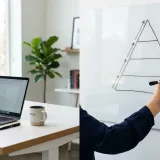 清潔な白いデスクに置かれたノートパソコン。その横で人がホワイトボードにピラミッド状の図解を丁寧に描いており、論理的な整理（ロジカルシンキング）を表現している。
