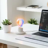清潔な白いデスクに置かれたノートパソコン。その横には明るく光る電球のオブジェがあり、内側から湧き出る意欲（モチベーション）を表現している。明るい自然光のプロフェッショナルな写真。