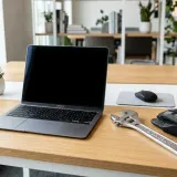 清潔な白いデスクに置かれたノートパソコン。その横には、プロフェッショナルな金属製のレンチと作業用の手袋が置かれ、現場で働く人々（ブルーカラー）を象徴している。明るい自然光のプロフェッショナルな写真。